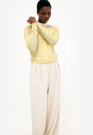 Kobieta w bladoróżowym kardiganie na guziki i luźnych kremowych plisowanych spodniach, stojąca z dłońmi złożonymi blisko twarzy.