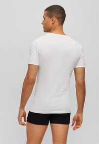 White fitted t-shirt with short sleeves, smooth fabric, and a crew neck, paired with black shorts. The back view shows the shirt's slight curve.