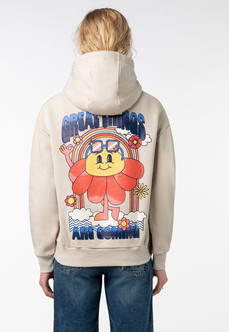 Lichtbeige hoodie met een grote afbeelding op de achterkant van een bloemenkarakter, een regenboog en de tekst "GROTE DINGEN KOMEN ERAAN."