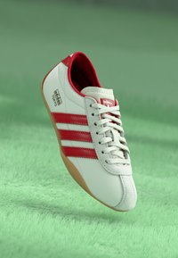 Fehér bőr sneaker piros díszítésekkel, három oldalsó csíkkal és gumi talppal. Kerek orrú és fűzős kialakítású.