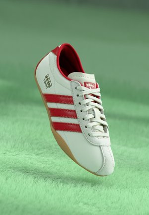 Sneaker in pelle bianca con accenti rossi, tre strisce laterali e suola in gomma naturale. Presenta punta rotonda e chiusura con lacci.