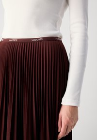 Femme portant un haut à manches longues côtelé blanc et une jupe plissée taille haute marron foncé avec une ceinture arborant le logo Lacoste.