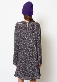 Blumenmuster-Langarmkleid in Dunkellila mit weißen und lilafarbenen Mustern; mit einem Schlüssel-ausschnitt und einem ausgestellten Saum. Getragen mit einer lila Mütze.