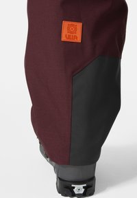 Bordeauxkleurige sneeuwbroeken met een zwarte onderzijde, voorzien van een oranje rubberen logo patch en stevige hardware voor een veilige pasvorm.