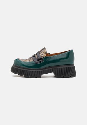 Mocassin vert foncé brillant avec semelle noire épaisse et détail texturé beige et noir sur l'avant supérieur, sur fond uni.