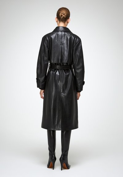 Donna con un chignon che indossa un trench coat in pelle nera con cintura e stivali in pelle nera al ginocchio, in piedi contro uno sfondo chiaro e uniforme.