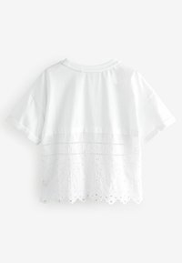 Vit t-shirt med ett lagerutformat design; övre halvan är i slät tyg, nedre halvan har spetsdetaljer med blommönster och scallopade kanter.