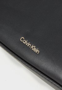 Schwarzes Lederportemonnaie mit glatter Textur, mit einem goldenen, auf der Vorderseite eingeprägten "Calvin Klein"-Logo. Kompaktes und schlankes Design.