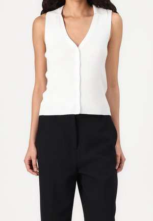 Femme portant un haut blanc sans manches côtelé avec boutons et col en V, et un pantalon noir taille haute sur un fond uni.