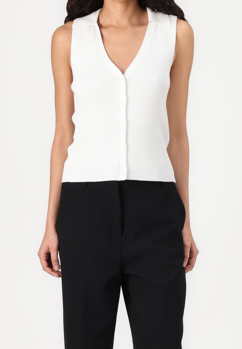 Femme portant un haut blanc sans manches côtelé avec boutons et col en V, et un pantalon noir taille haute sur un fond uni.