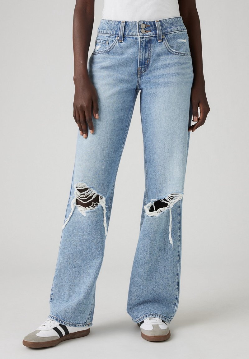 Personne portant un jean bleu clair à jambes larges avec de grandes déchirures aux genoux et des baskets blanches à bandes noires.