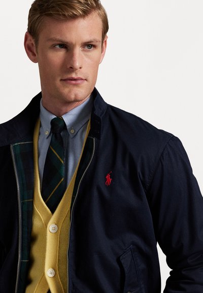 Marineblaues Jacket mit rot gesticktem Logo, über einem gelben Cardigan getragen. Ein hellblaues Hemd mit Knöpfen und eine gestreifte Krawatte runden das Outfit ab.