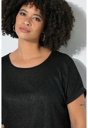Femme aux cheveux bouclés, boucles d'oreilles en or en forme d'anneaux et haut court noir scintillant, debout devant un mur gris clair.