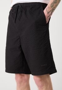 Schwarze Shorts aus strukturiertem Stoff, mit einem elastischen Bund und Kordelzug sowie seitlichen Taschen. Logodetail ist sichtbar.