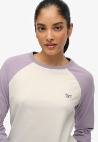 Top de manga raglán en crema con mangas lila claras. La suave tela de algodón presenta un pequeño logo bordado de "Super Dry" en el pecho.