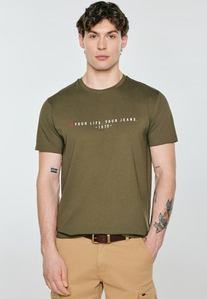 Młody mężczyzna z kręconymi włosami nosi oliwkowo zieloną koszulkę z napisem „YOUR LIFE. YOUR JEANS. -1979-” oraz beżowe spodnie z tatuażami na ramionach.