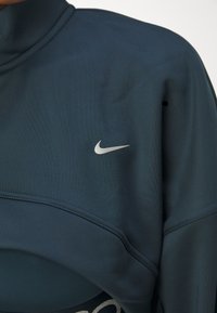 Mornarijevo plavi sportski top s visokim ovratnikom, izrađen od glatkog materijala, sadrži srebrni Nike logotip na lijevoj strani grudi i stilizirano šivanje.