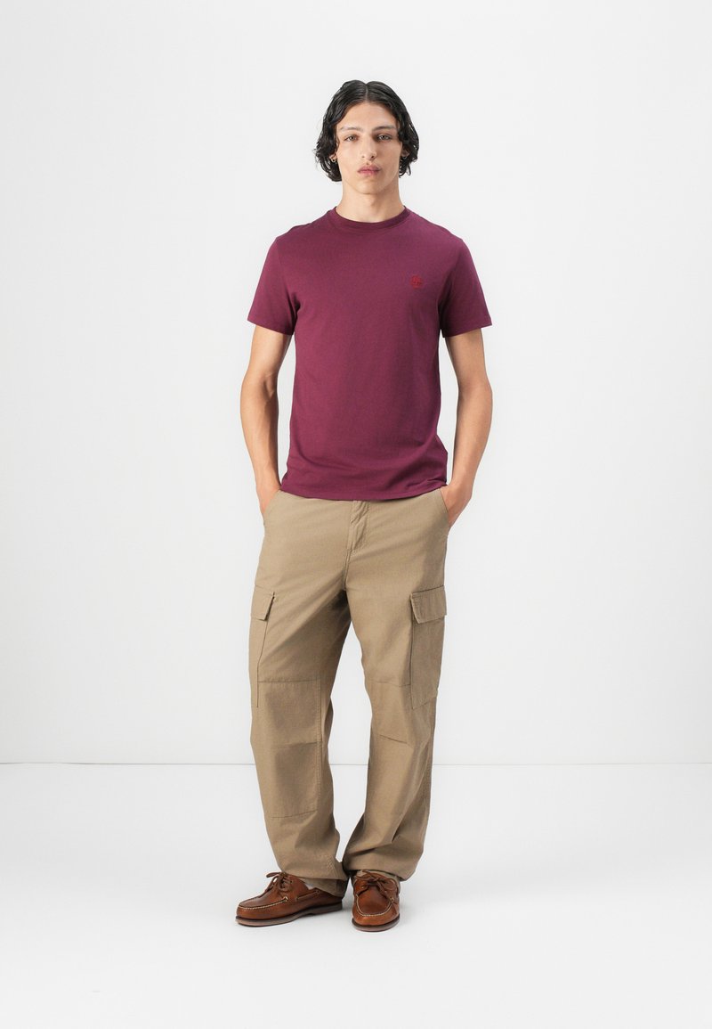 T-shirt corto bordeaux con un logo discreto, abbinato a pantaloni cargo beige con tasche laterali e scarpe da barca marroni.