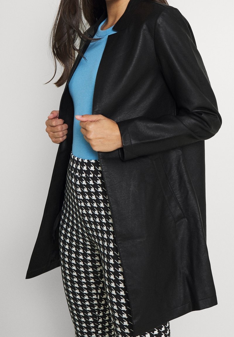 Abrigo largo de cuero sintético negro con un diseño elegante, combinado con una blusa azul y pantalones con patrón de pata de gallo en blanco y negro.