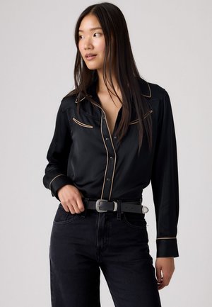 Camicia nera in satin con piping beige, maniche lunghe e colletto classico, abbinata a jeans scuri e una cintura nera.