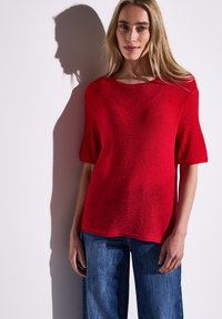 Roter kurzärmeliger Strickpullover mit Rundhalsausschnitt, der einen strukturierten Stoff und eine lockere Passform aufweist, kombiniert mit blauen weit geschnittenen Jeans.