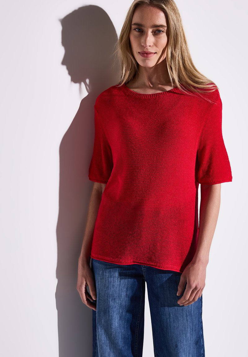 Roter kurzärmeliger Strickpullover mit Rundhalsausschnitt, der einen strukturierten Stoff und eine lockere Passform aufweist, kombiniert mit blauen weit geschnittenen Jeans.