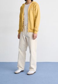 Gul zip-up hoodie, vit t-shirt, ljusbeige byxor och vita sneakers med bruna detaljer. Mjuk tyg, avslappnad passform, casual design.