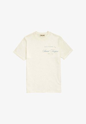 T-shirt beige à manches courtes avec col rond et texte bleu sur la poitrine indiquant « Welcome to Saint Tropez Été 1984 ».