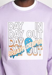 Lavendfärgad sweatshirt med en stor vit grafik och svart text som säger "DAY IN DAY OUT" och blå accenter, inklusive frasen "BREATH DEEPER."
