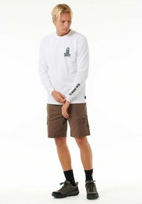 Camisa blanca de manga larga con un pequeño logo azul en el pecho y texto negro en la manga, combinada con pantalones cortos de carga marrones y zapatos oscuros.