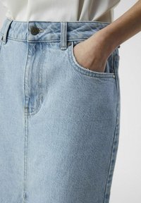 Jupe en jean bleu clair à taille haute, avec poche frontale et détail de bouton en laiton. Tissu texturé et bord effiloché au niveau de la poche.