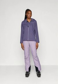 Sieviete stāv ar violetu ar rāvējslēdzēju sabojātu jaku, gaiši violetām biksēm un melniem slēpošanas zābakiem pret vienkāršu balto fonu.