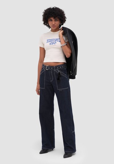 Femme aux cheveux bouclés portant un t-shirt graphique blanc court, un jean large foncé avec ceinture, tenant une veste noire sur une épaule.