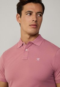 Růžová polo košile s texturovanou tkaninou, klasickým límcem, dvěma knoflíky a malým bílým logem na levé hrudi.
