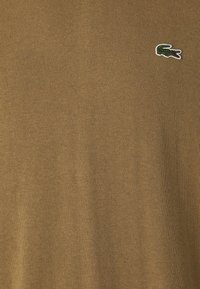Khaki Strickpullover mit einem kleinen gestickten grünen Krokodil-Logo auf der linken Brust. Die Textur ist glatt und gleichmäßig, mit einem gerippten Finish.