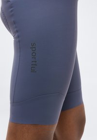 Κοντινό πλάνο του άνω μέρους των ποδιών ενός ατόμου που φοράει στενά, γκρι-μπλε σορτς ποδηλασίας με το λογότυπο «Sportful» στον αριστερό μηρό.