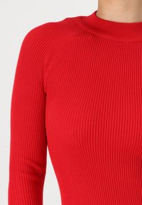 Rød ribbestrikket topp med høy krage og lange ermer, med en tettsittende silhuett og teksturert mønster.