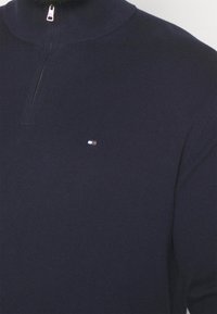 Pull-over marine avec un col haut, fermeture éclair demi-longue et texture côtelée. Porte un petit logo sur la poitrine gauche.
