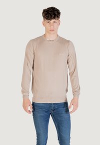 Maglione beige lavorato a maglia con collo rotondo, maniche lunghe e vita aderente. Dettaglio logo sottile sul petto. Abbinato a jeans blu.
