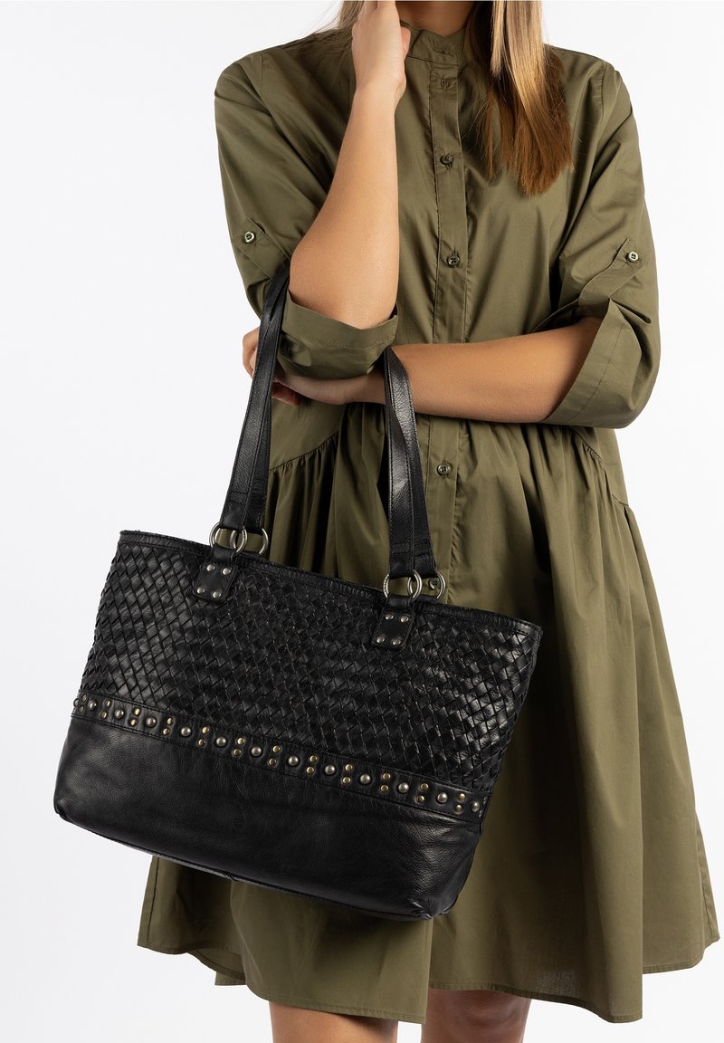 Femme en robe verte tenant un sac à main en cuir noir tissé avec des clous en métal et des doubles bretelles sur fond blanc.