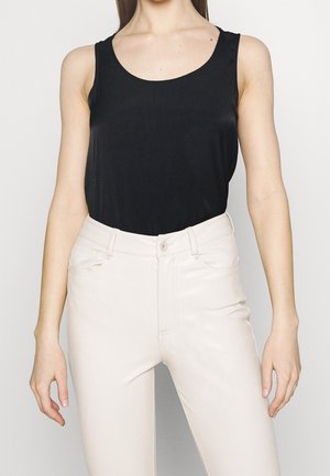 Vrouw met een zwart mouwloos topje dat is ingestopt in een witte broek met hoge taille, staand met ontspannen armen langs het lichaam tegen een effen achtergrond.