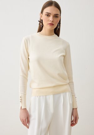 Mujer con cabello largo castaño lleva un suéter de manga larga color crema y pantalones blancos plisados, de pie contra un fondo claro liso.