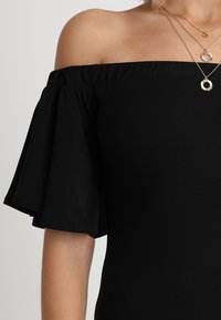 Haut noir à épaules dénudées avec manches courtes évasées. Tissu doux au toucher lisse et design minimaliste. Colliers dorés superposés pour plus de détails.