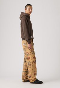 Sudadera con capucha marrón y bolsillo delantero, combinada con pantalones amarillos que presentan gráficos impresos de estilo vintage y un corte holgado. Calzado negro.