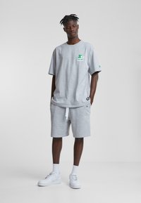 T-shirt gris en coton à manches courtes avec un logo vert, associé à un short gris assorti avec cordon de serrage et patch logo, porté avec des baskets blanches.