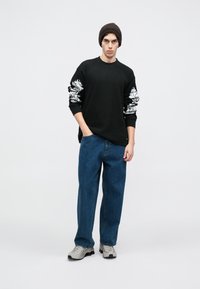 Μαύρο μπλουζάκι με μακριά μανίκια και λευκά γραφικά σχέδια στα μανίκια, συνδυασμένο με γαλάζια φαρδιά τζιν και γκρίζα sneakers. Το μοντέλο φοράει ένα καφέ πλεκτό σκουφί.