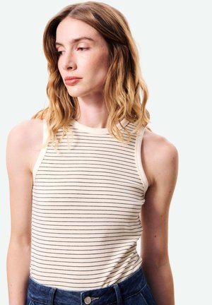 Jeune femme aux cheveux blonds mi-longs portant un haut sans manches rayé blanc et noir et un jean bleu, regardant vers sa gauche.