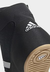 Gros plan sur le talon d'une chaussure Adidas noire avec logo blanc et semelle en caoutchouc beige montrant la marque Adidas.