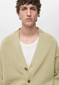 Beige stickad cardigan med djup V-ringning, prydd med två bruna knappar. Bärs över en vit ribbad t-shirt. Mjuk textur och avslappnad passform.