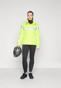 High-visibility yellow rain jacket with silver reflective stripes, paired with black fitted leggings and sneakers. Cycling helmet held in one hand.